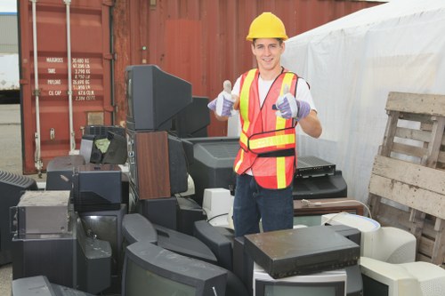 A young male worker wearing a yellow safety helmet, high-visibility vest with orange and yellow stripes, and purple work gloves standing outdoors in an industrial or storage yard setting. He is smiling and giving a thumbs-up gesture with his right hand while holding a plastic bottle in his left hand. Surrounding him are various discarded electronic devices such as old televisions, monitors, and other obsolete electronic equipment, stacked haphazardly on a paved surface. In the background, there is a large shipping container and a wooden pallet leaning against a white tarpaulin covering something. The image visually relates to rubbish removal services, particularly for electronic waste, and is associated with Business Waste Removal Neasden, serving the local area near NW10 postcode. The scene captures a professional approach to clearing and recycling bulky or electronic rubbish, emphasizing the importance of proper waste disposal and de-cluttering in a commercial or residential context in the Neasden area.