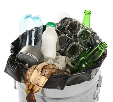 A close-up image of a metal rubbish bin lined with a black plastic bag, filled with various waste items including a transparent plastic water bottle with a blue cap, a white plastic bottle with a green lid, several used black plastic food containers, a crushed aluminum can, and a piece of brown, weathered wood or cardboard. The waste is piled visibly and appears to be outdoor or on a driveway setting, with some items leaning against the edge of the bin, demonstrating typical household or business rubbish for collection in the Neasden area. The background is neutral and unobstructed, focusing solely on the refuse inside the bin, aligning with rubbish removal services provided by Business Waste Removal Neasden as part of their local waste management efforts.
