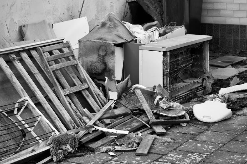 A collection of discarded household items and waste situated outdoors on a paved surface. The scene features a wooden garden chair with slatted legs and a slatted backrest, leaning against a nearby wa