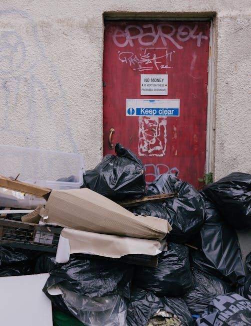 A collection of discarded household items and waste situated outdoors on a paved surface. The scene features a wooden garden chair with slatted legs and a slatted backrest, leaning against a nearby wa