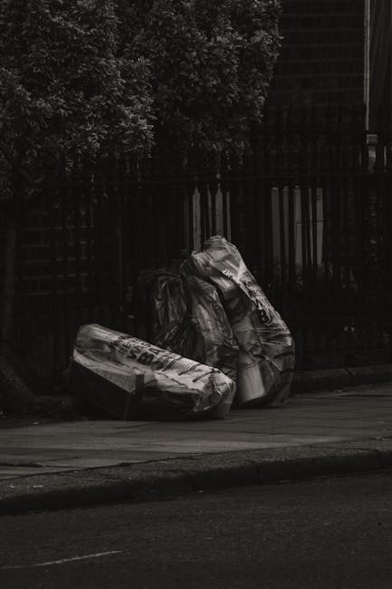 A narrow urban alleyway cluttered with large piles of mixed waste and discarded materials. In the foreground, a large, weathered black and gray garbage bag is secured onto a trolley, ready for collect