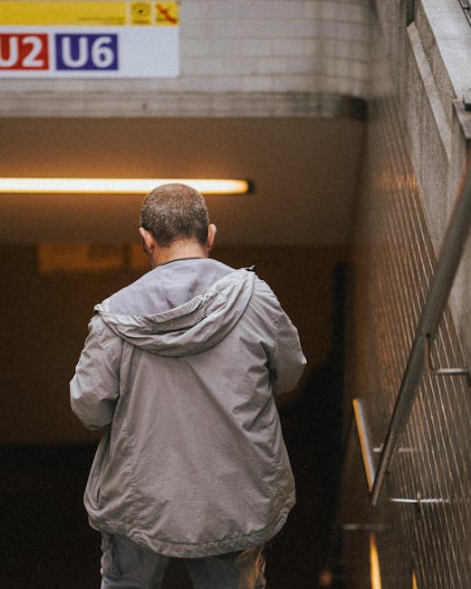 A person with short hair dressed in a light grey hooded jacket is walking up a staircase in an underground setting, seen from behind. The staircase has a metal handrail on the right side and steps tha